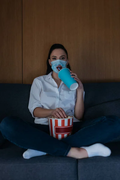 Junge Frau in medizinischer Maske mit Loch sitzt mit überkreuzten Beinen auf dem Sofa, trinkt Limo, isst Popcorn und schaut fern — Stockfoto