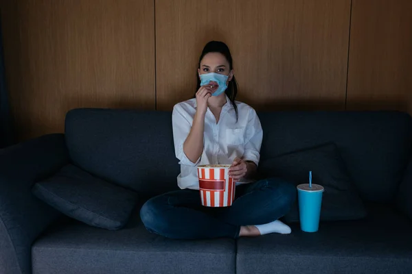 Junge Frau in medizinischer Maske mit Loch sitzt mit überkreuzten Beinen auf dem Sofa, schaut fern und isst Popcorn — Stockfoto