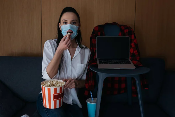 Jovem mulher em máscara médica com buraco comendo pipocas enquanto assiste tv no sofá perto de laptop com tela em branco na cadeira — Fotografia de Stock