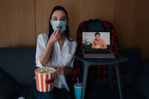 Молода жінка в медичній масці з отвором їсть попкорн біля ноутбука з усміхненим азіатським хлопцем на екрані — стокове фото