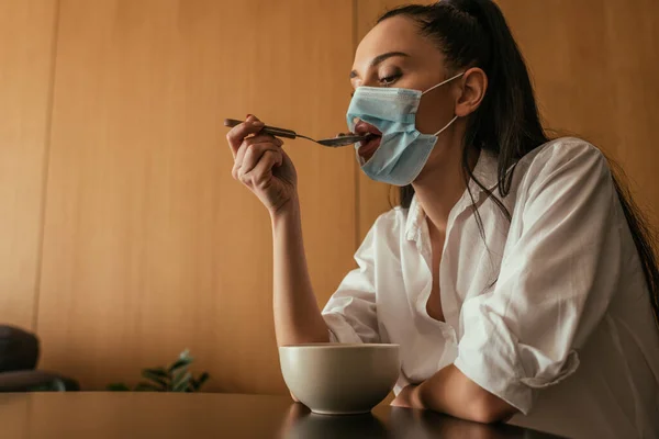 Mujer joven en máscara médica con agujero desayunando en casa - foto de stock