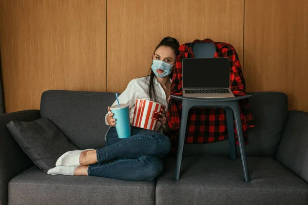 Mujer joven en máscara médica con agujero sosteniendo palomitas de maíz y refrescos cerca de la computadora portátil con pantalla en blanco en la silla - foto de stock