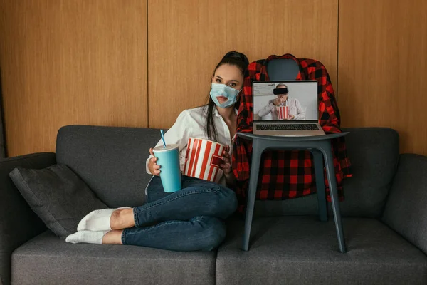Jovem mulher em máscara médica com buraco segurando pipoca e refrigerante perto de laptop com namorado no fone de ouvido vr na tela — Fotografia de Stock