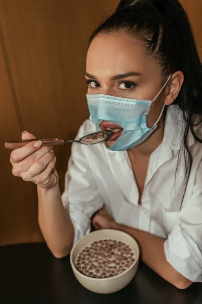 Jeune femme en masque médical avec trou pour la bouche petit déjeuner et regarder la caméra — Photo de stock