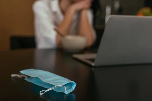 Селективный фокус медицинской маски на столе рядом с женщиной и ноутбук на заднем плане — стоковое фото
