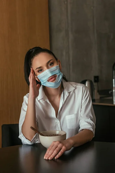Femme déprimée dans un masque médical avec trou regardant la caméra tout en étant assis près du bol avec le petit déjeuner — Photo de stock