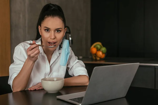 Mujer joven desayunando y mirando a la cámara cerca del ordenador portátil en la cocina - foto de stock