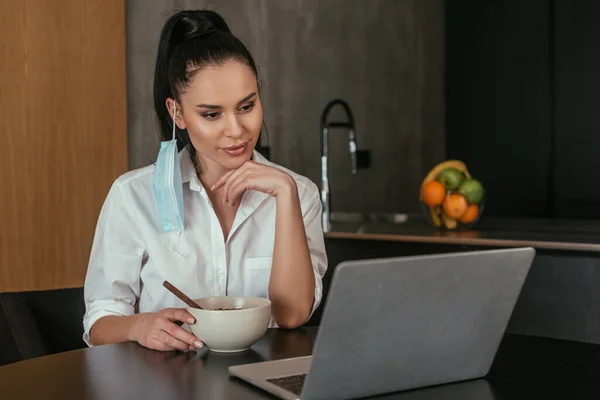 Menina sorrindo olhando para laptop enquanto sentado na mesa perto de tigela com café da manhã — Fotografia de Stock