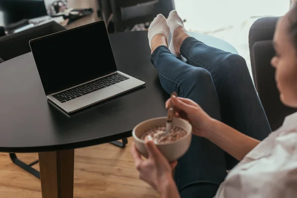 Selective focus of girl holding bowl with breakfast while looking at laptop with blank screen — Stock Photo