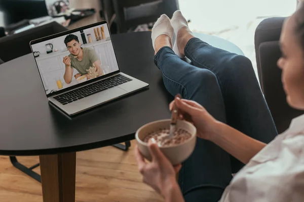 Селективное внимание девушки, держащей миску с хрустами во время видеочата с веселым азиатским парнем, завтракающим — стоковое фото