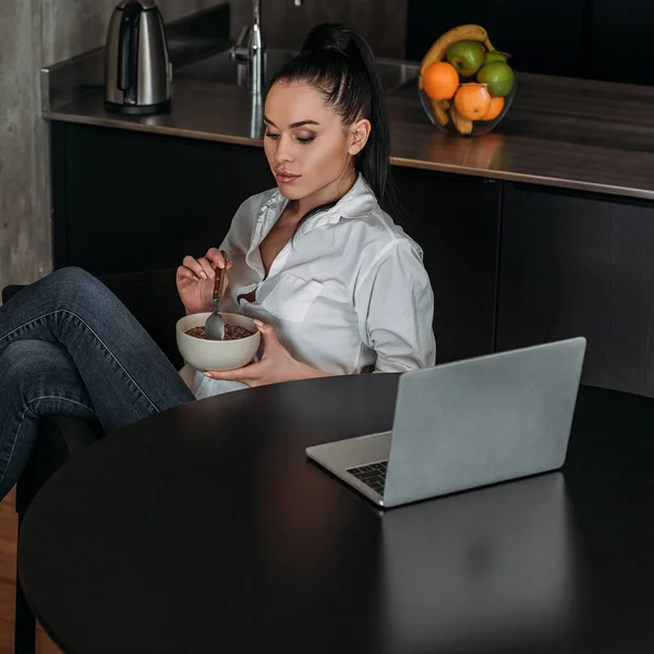Вдумчивая женщина держит миску с завтраком, сидя рядом с ноутбуком — стоковое фото