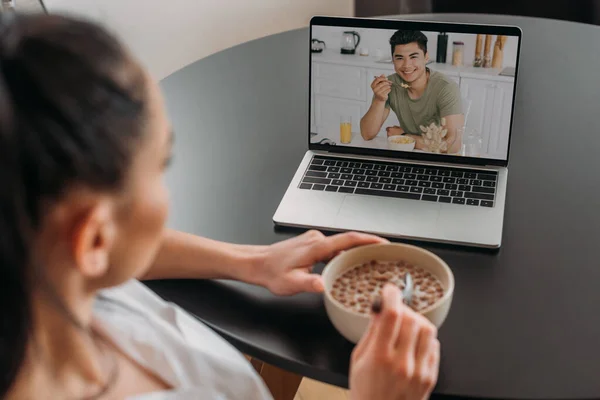 Selective focus of girl holding bowl with muesli during video chat with smiling asian boyfriend having breakfast — Stock Photo