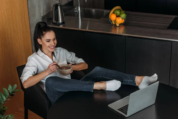 Cheerful woman having breakfast while sitting with legs on table and looking at laptop — Stock Photo