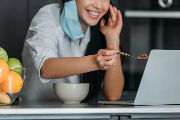 Vista recortada de la mujer sonriente sosteniendo cuchara con muesli cerca de la computadora portátil en la cocina - foto de stock