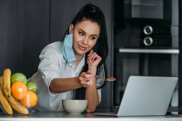 Sonriente mujer sosteniendo cuchara con muesli cerca de la computadora portátil en la cocina - foto de stock