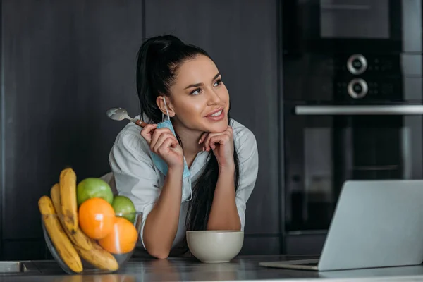 Sorridente, donna sognante sorridente mentre tiene cucchiaio vicino al computer portatile, ciotola e frutta in cucina — Foto stock