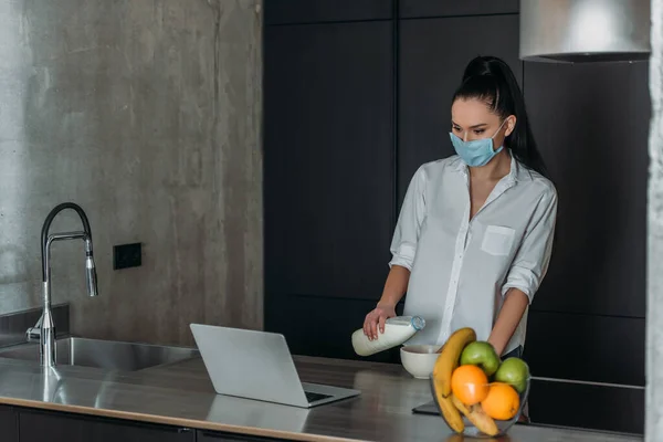 Jeune femme dans le masque médical verser du lait dans le bol tout en regardant ordinateur portable dans la cuisine — Photo de stock