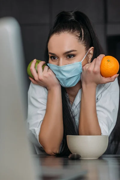 Selective focus of young woman in protective mask holding orange and apple during video chat on laptop — Stock Photo