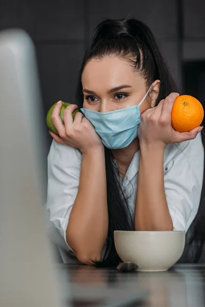 Enfoque selectivo de la mujer joven en la máscara protectora celebración de naranja y manzana durante el chat de vídeo en el ordenador portátil - foto de stock