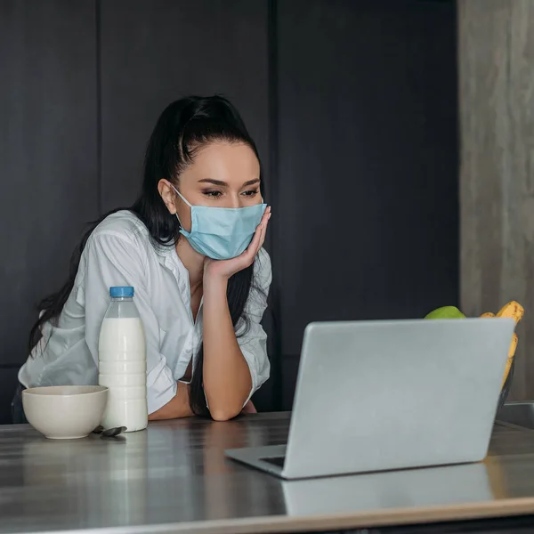 Young woman in medical mask looking at laptop near bottle of milk in kitchen — Stock Photo