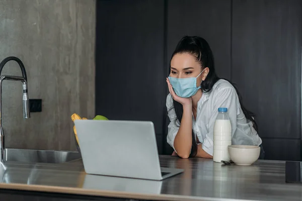 Young woman in protective mask looking at laptop near bottle of milk in kitchen — Stock Photo