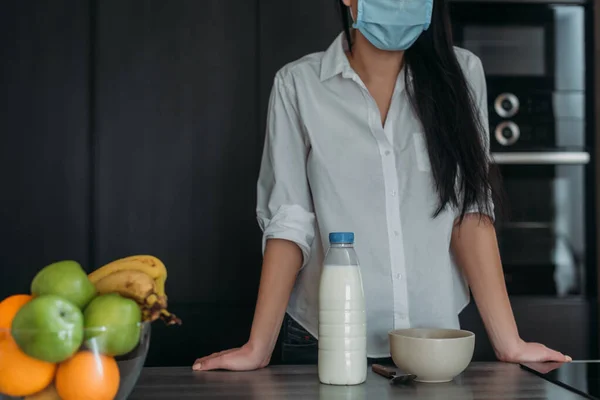 Cropped view of woman in protective mask near bottle of milk, bowl and fresh fruits in kitchen — Stock Photo