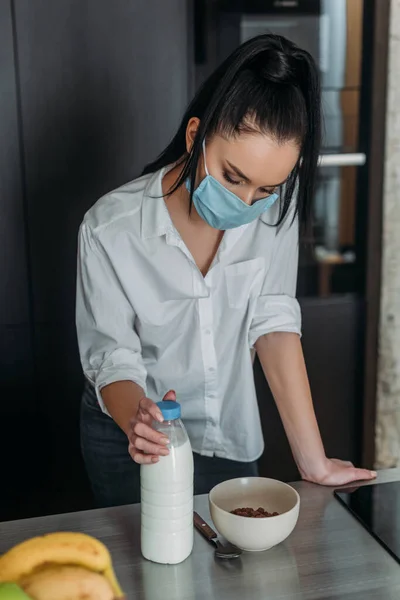 Depressed woman in medical mask standing with bowed head while touching bottle of milk in kitchen — Stock Photo