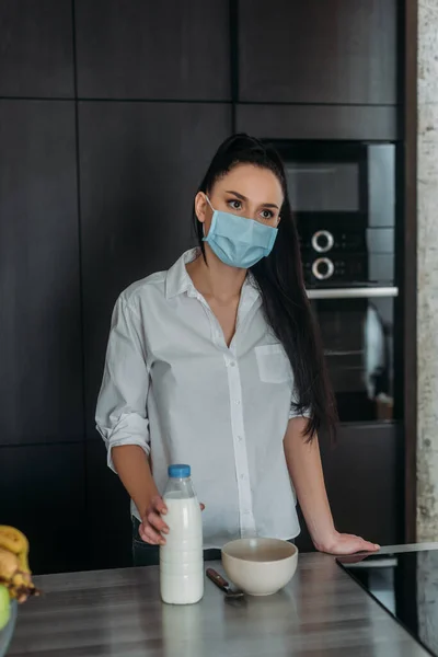 Femme triste dans le masque médical regardant loin tout en touchant bouteille de lait dans la cuisine — Photo de stock