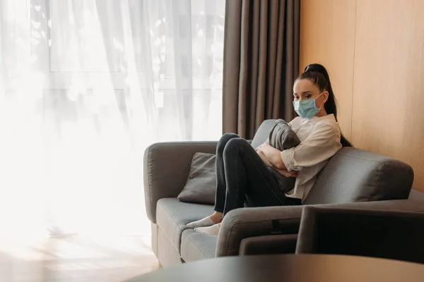 Mujer joven disgustada en máscara protectora sentada en el sofá y abrazando la almohada - foto de stock