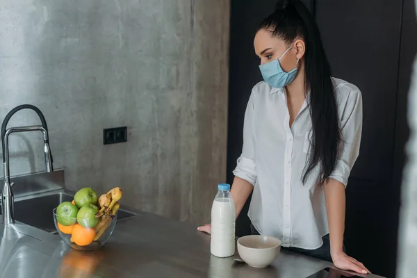 Femme déprimée dans un masque médical près de bouteille de lait, fruits frais et bol dans la cuisine — Photo de stock