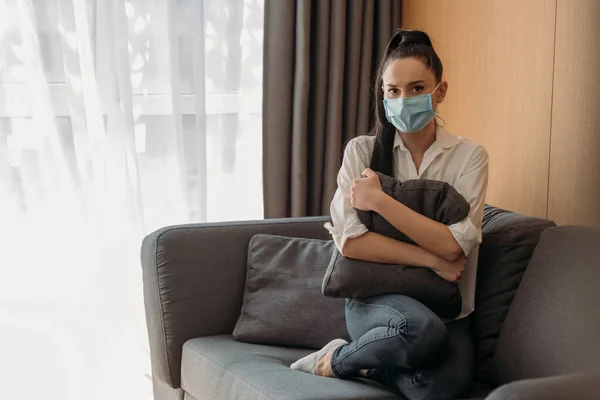 Depresso giovane donna in maschera medica guardando la fotocamera mentre seduto sul divano e abbracciando cuscino — Foto stock