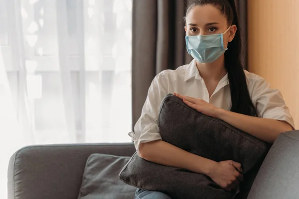 Triste jovem mulher em máscara médica olhando para longe enquanto sentado no sofá e abraçando travesseiro — Fotografia de Stock