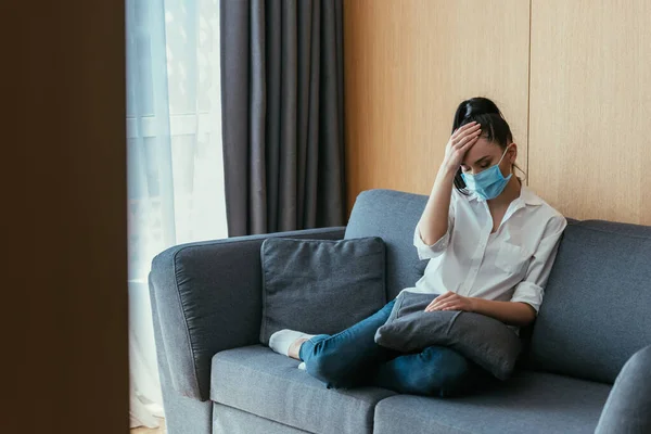 Mujer joven deprimida en máscara médica tocando la frente mientras está sentado en el sofá con los ojos cerrados - foto de stock