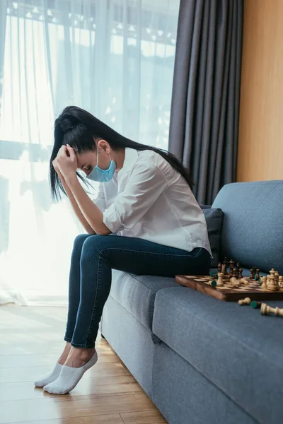Depressed woman in protective mask sitting on sofa near chessboard with bowed head — Stock Photo