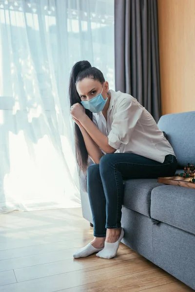 En colère, femme déprimée dans le masque médical assis sur le canapé et regardant la caméra — Photo de stock