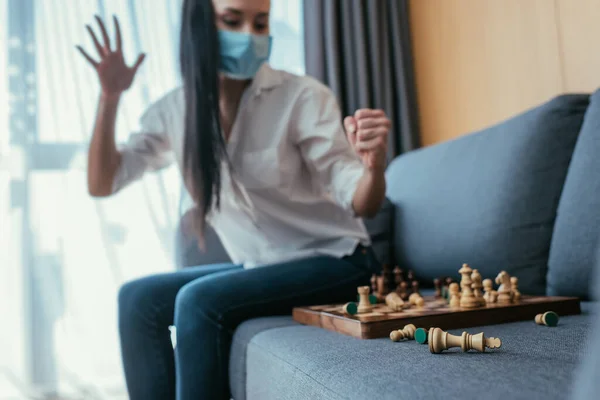 Enfoque selectivo de enojado, mujer deprimida en máscara protectora chocar tablero de ajedrez mientras está sentado en el sofá - foto de stock