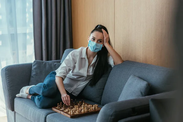 Enfoque selectivo de la mujer deprimida en máscara protectora sentado en el sofá cerca de tablero de ajedrez - foto de stock