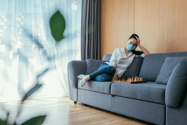 Вибірковий фокус сумної жінки в медичній масці, що сидить біля шахівниці на дивані — стокове фото
