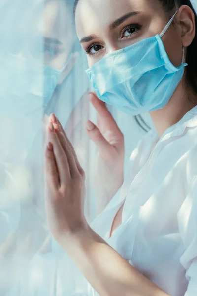 Triste fille dans le masque médical touchant le verre de fenêtre et détournant les yeux — Photo de stock