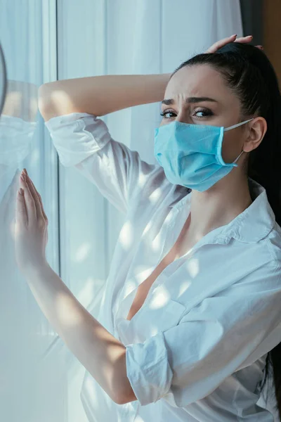 Menina deprimida em máscara médica tocando cabeça enquanto estava perto da janela em casa — Fotografia de Stock