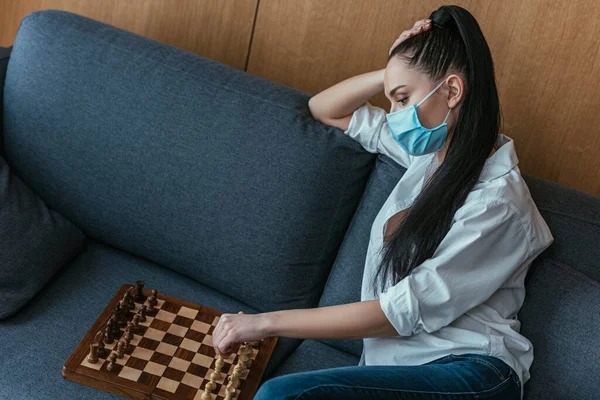 Sad woman in medical mask sitting on sofa near chessboard — Stock Photo