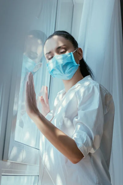 Niedrigwinkel-Ansicht eines depressiven Mädchens in medizinischer Maske, das sich zu Hause mit geschlossenen Augen am Fenster lehnt — Stockfoto