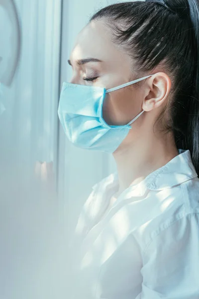 Foco seletivo da mulher chateada na máscara médica que está perto da janela com olhos fechados — Fotografia de Stock