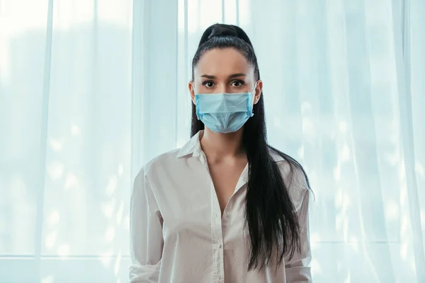 Ernstes Mädchen mit medizinischer Maske, das in die Kamera schaut, während es zu Hause am Fenster steht — Stockfoto