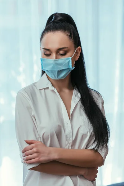 Депрессивная девушка в медицинской маске стоит со скрещенными руками дома — стоковое фото