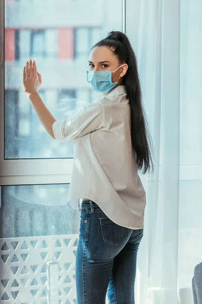 Сумна дівчина в медичній масці торкається віконного скла і дивиться на камеру — стокове фото