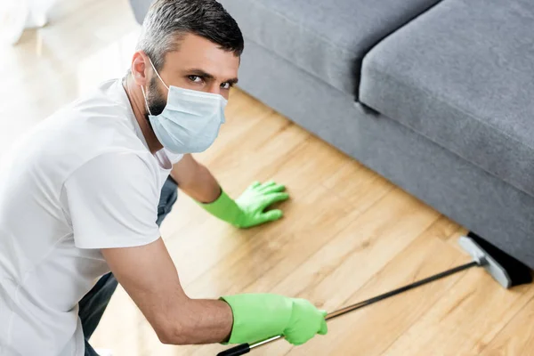 Homme en masque médical regardant la caméra tout en nettoyant le sol dans le salon — Photo de stock