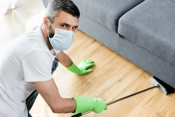 Homem em máscara médica olhando para a câmera enquanto limpava o chão na sala de estar — Fotografia de Stock