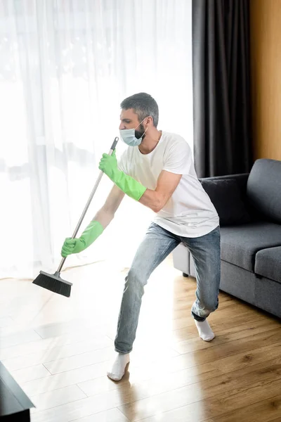 Mann mit medizinischer Maske und Gummihandschuhen hält Besen beim Fußbodenputzen — Stockfoto