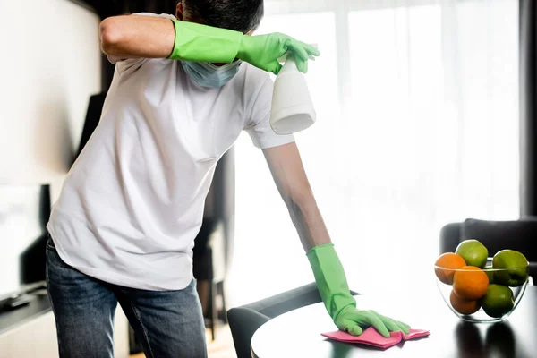 Mann mit medizinischer Maske und Gummihandschuhen zu Hause Putztisch mit Lappen und Spülmittel — Stockfoto